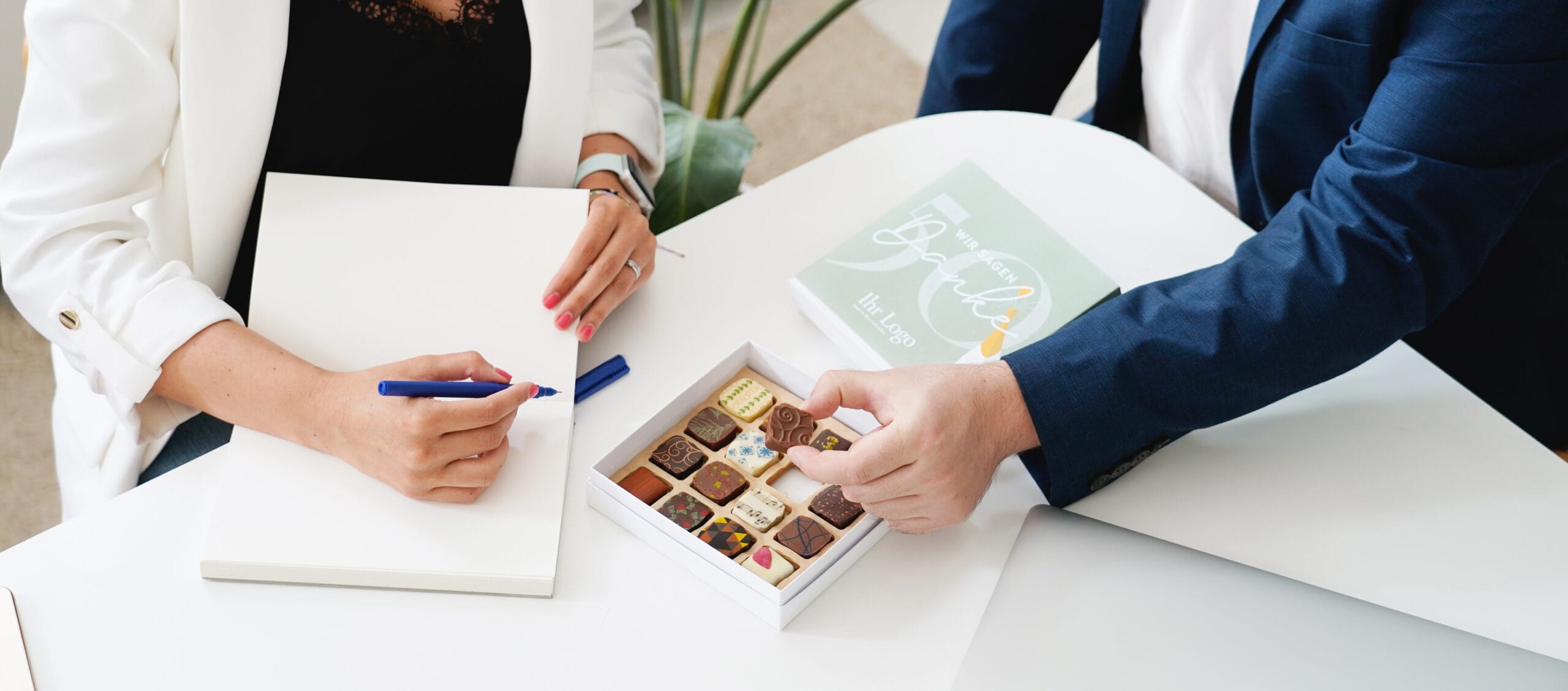 Zwei Personen sitzen an einem Büro Tisch. Man sieht nur die Hände. Eine Frau schreibt in ein Notizbuch, der Mann greift in eine geöffnete Pralinenbox mit bunten Pralinen.