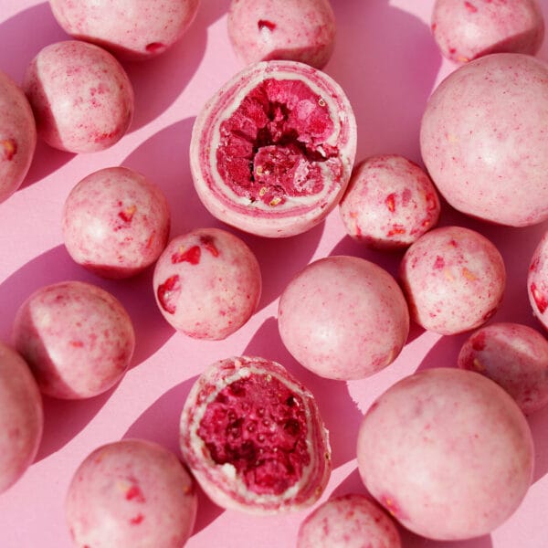 Himbeeren in weißer Schokolade. Eine ist aufgeschnitten, sodass die gefriergetrocknete Himbeere mit der Schokoladenhülle sichtbar ist