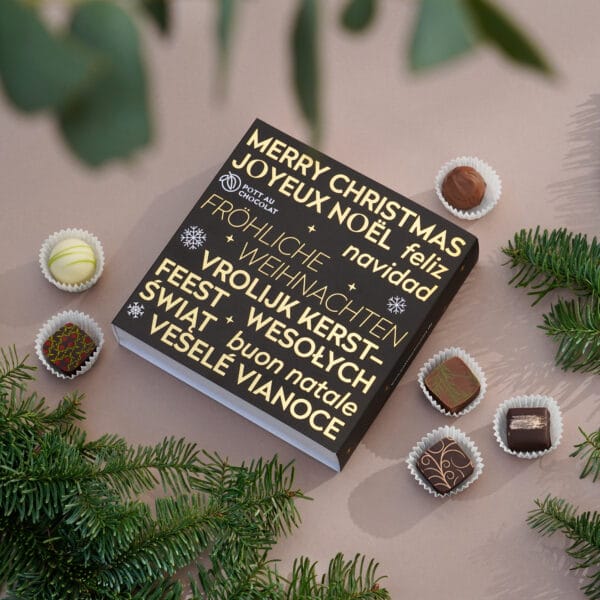 Quadratische Pralinenschachtel mit schwarzer Banderole auf beigem Hintergrund. 'Fröhliche Weihnachten' in gold geprägt in verschiedenen Sprachen ist das Motiv der Banderole. Daneben sind einige bunt dekorierte Pralinen und grüne Zweige.