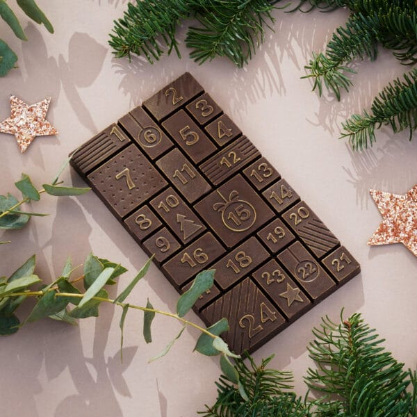 Schokoladentafel in dunkler Schokolade in Adventskalenderform gegossen. Die einzelnen Stückchen der Schokolade zeigen die Zahlen 1-24. Die Schokolade glänzt leicht gold und liegt auf beigem Hintergrund. Daneben verschiedene Zweige und Sterne.