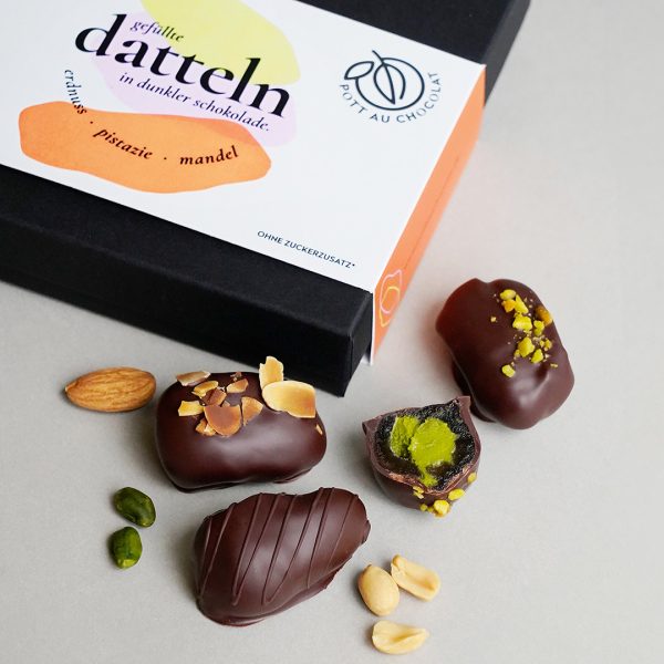 3 Datteln überzogen mit dunkler Schokolade und Nuss Dekor liegen vor der verschlossenen Dattel Verpackung. Eine schwarze Box mit bunter Banderole.