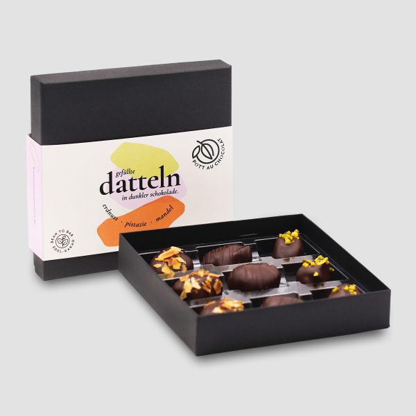Verpackung der Datteln - eine schwarze Box mit heller Banderole mit den Farben Beige, rosa, grün und orange. Davor die Schachtel geöffnet mit 9 Datteln.