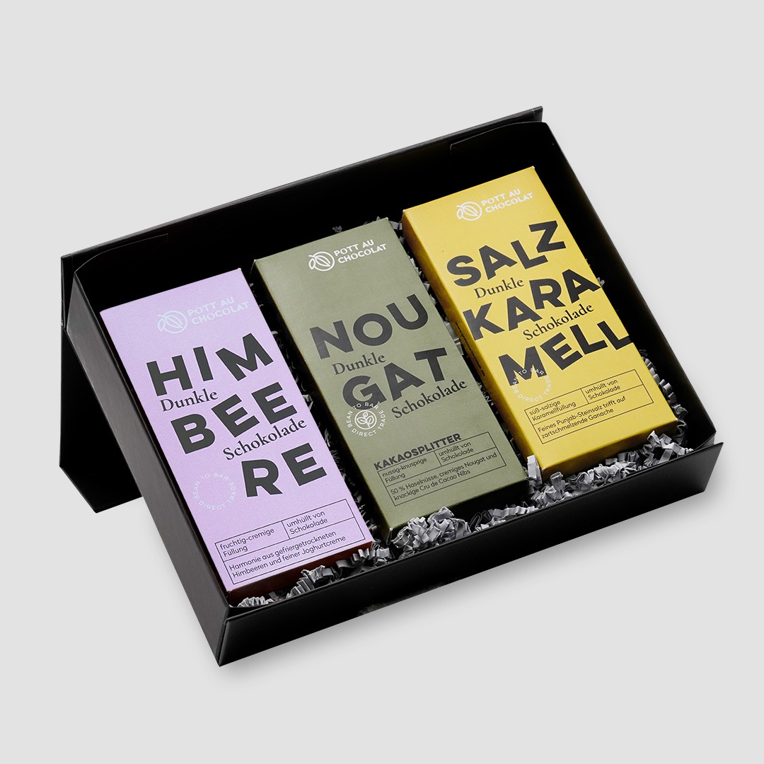 Geöffnete schwarze Geschenkbox mit drei rechteckigen Schokoladentafeln nebeneinander. Die Verpackungen sind einfarbig in rosa, dunkelgrün und gelbgrün mit schwarzer Typografie. Sorten: Himbeere, Nougat Kakaosplitter und Salzkaramell.