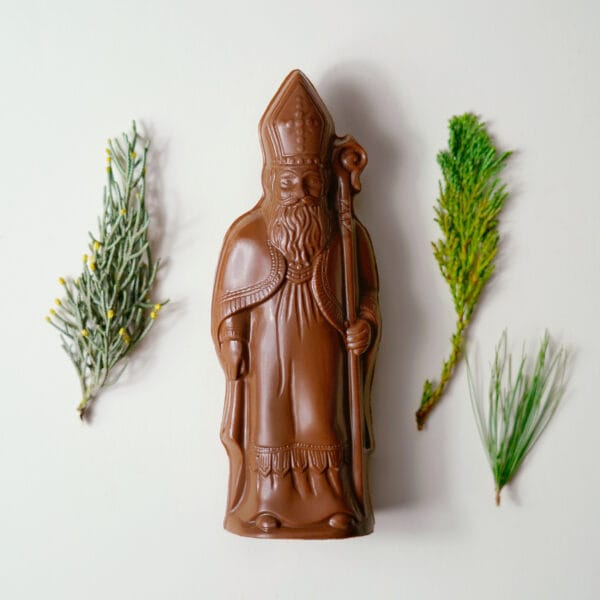 Ein klassischer Nikolaus in Milchschokolade liegt neben verschiedenen Zweigen auf hellgrauem Hintergrund.
