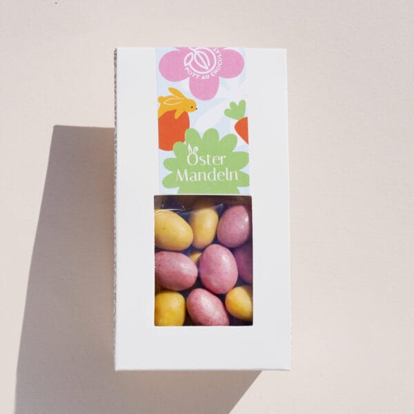 Verpackung mit Fenster, durch das man das Produkt sehen kann: Mandeln umhüllt von Schokolade in Gelb und Rosa. Die Verpackung ist im Osterdesign gestaltet, mit der Aufschrift 'Ostermandeln'. Farben des Designs: Rosa, Orange und Hellgrün