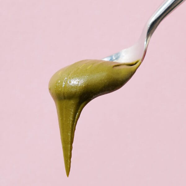 grüner Pistazien Aufstrich fliesst von einem Löffel vor rosa Hintergrund