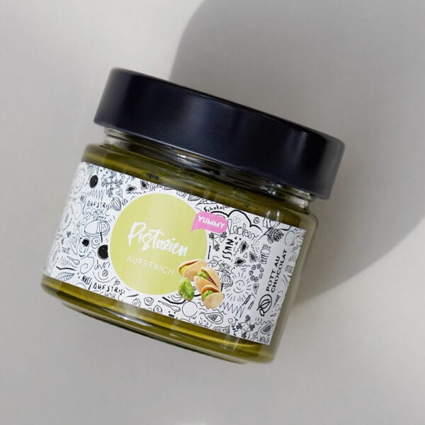 Schräge Ansicht eines Glases mit breitem schwarzen Deckel mit Pistazien-Aufstrich. Das Etikett ist Weiß mit einem grünen Kreis in der Mitte mit der Aufschrift. Drumherum ganz viele kleine Zeichnungen in Schwarz.