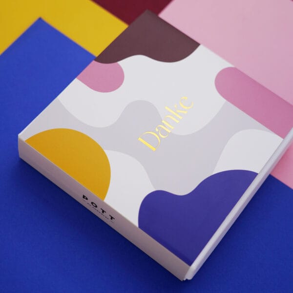 Geschlossene Pralinenbox liegend auf bunten Papieren in den Farben des Box-Designs