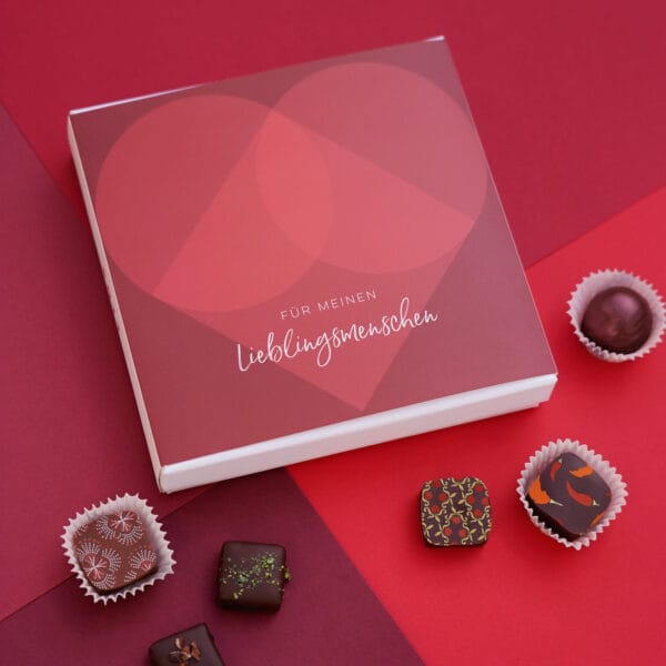 Pralinenbox 'Lieblingsmensch' in weiß mit roter Banderole mit großem roten Herz, davor ein paar bunt dekorierte Pralinen auf rotem Hintergrund.
