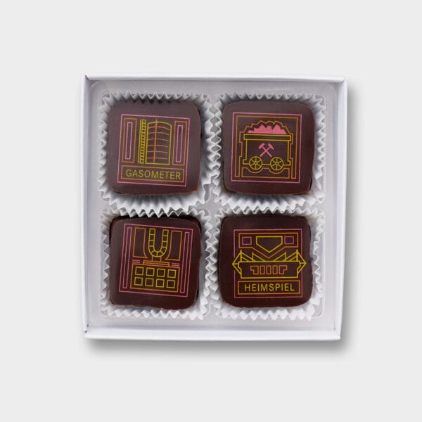 4 Pralinen in einer geöffneten weißen Pralinenschachtel. Die Pralinen in dunkler Schokolade zeigen 4 Motive aus dem Ruhrgebiet: das Gasometer, einen Grubenwagen, das Dortmunder U und das Dortmunder Stadion mit der Aufschrift 'Heimspiel'. Die Motive sind in den Farben gelb, orange und pink,