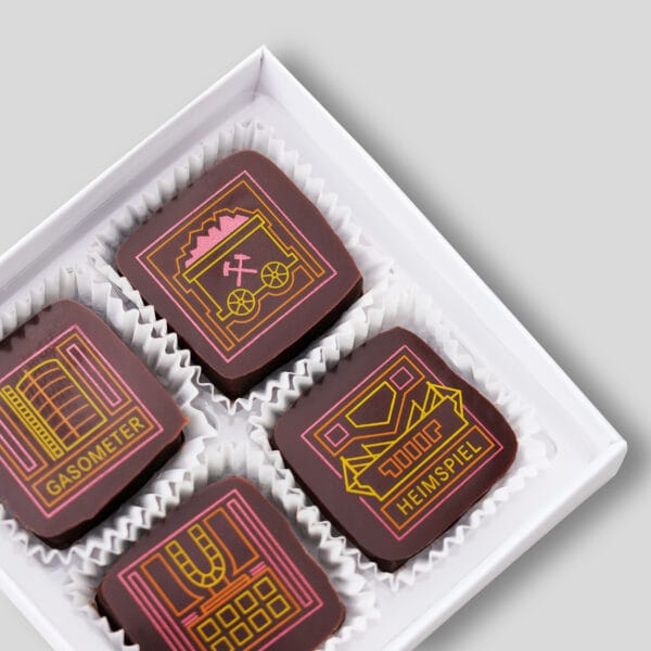 4 Pralinen in einer geöffneten weißen Pralinenschachtel. Die Pralinen in dunkler Schokolade zeigen 4 Motive aus dem Ruhrgebiet: das Gasometer, einen Grubenwagen, das Dortmunder U und das Dortmunder Stadion mit der Aufschrift 'Heimspiel'. Die Motive sind in den Farben gelb, orange und pink,