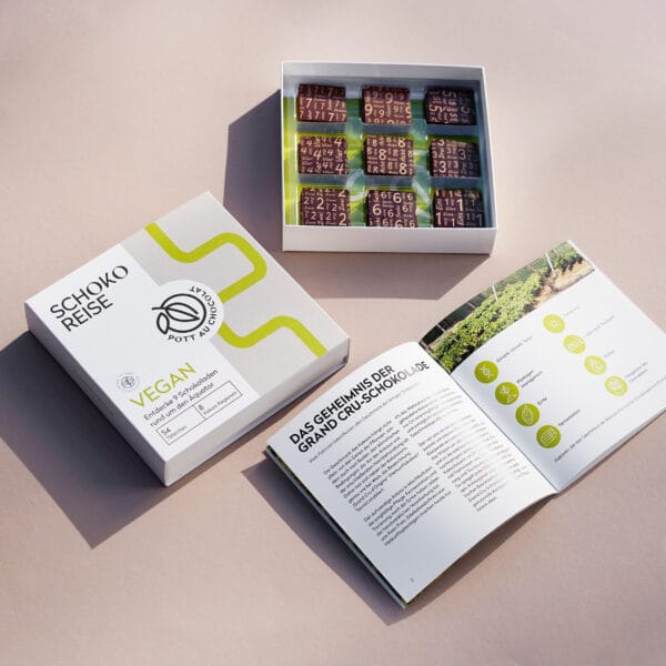 Schoko-Reise' Box mit 9 verschiedenen Probiertafeln auf hellbraunem Untergrund . Zu sehen ist die Banderole der Box in Weiss und Grau, die offene Box mit den Täfelchen und eine aufgeklappte Broschüre mit interessanten Informationen zu den Tafeln