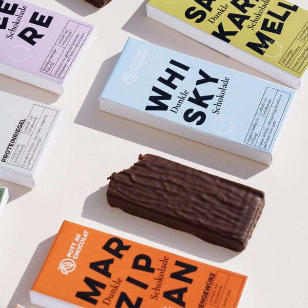 Mehrere Verpackungen der gefüllten Tafeln – Sorten wie Marzipan, Whisky, Salzkaramell, Himbeere und mehr. Die Verpackungen sind bunt gestaltet in Orange, Hellblau, Gelb und Rosa. Zwischen den Verpackungen liegt eine Tafel der gefüllten Schokolade