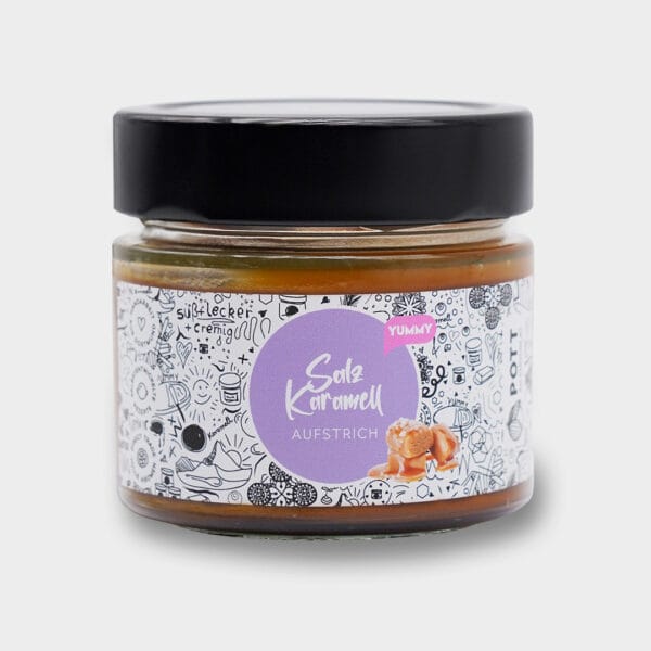 Frontansicht Salz-Karamell-Aufstrich in einem Glas mit breitem schwarzen Deckel. Das Glas hat vorne ein weißen Etikett versehen. In der Mitte ein Lila Kreis mit der Aufschrift. Davor Karamellbissen. Drumherum ganz viele kleine Zeichnungen in Schwarz.