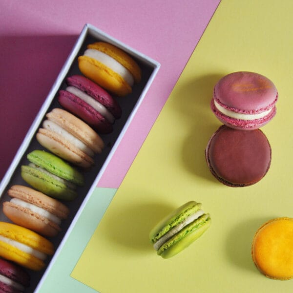 einreihige geöffnete Macaronbox von oben auf buntem Tonerpapier in Gelb, Lila und Mint. Auf der rechten Seite liegen vier einzelne Macarons eines davon hochkant