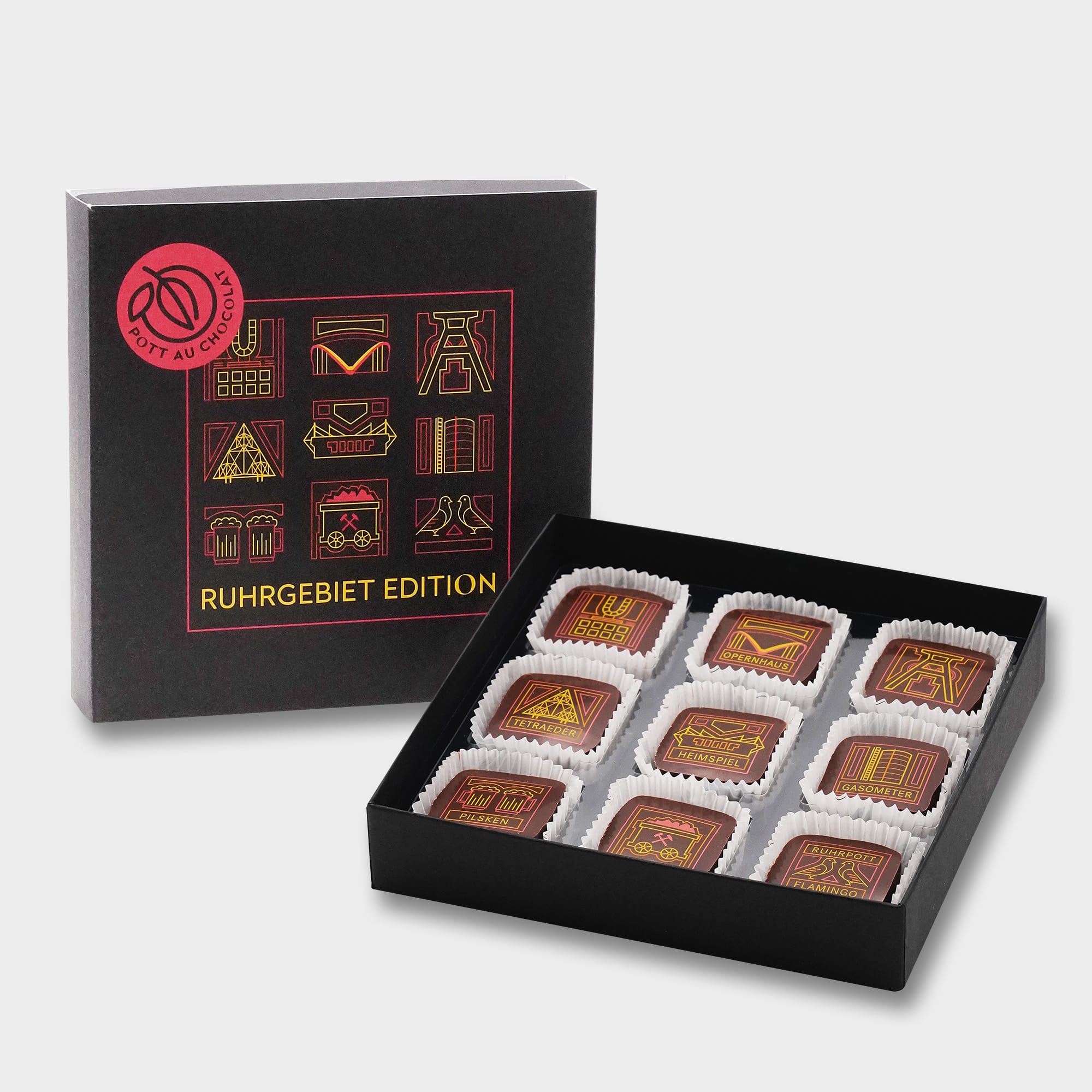 Schwarze Pralinenbox die vorne die Zeichnungen der Ruhrgebiets-Pralinen zeigt über dem Titel RUHRGEBIET EDITION in Grossbuchstaben in Gelb Orange und Rot auf schwarezm Grund. Davor liegt die geöffnete Box mit den Pralinen mit Zeichnungen von unterschiedlichen Wahrzeichen aus dem Ruhrpott wie Dortmunder U, der Doppelförderturm, eine Kohlenlohre, das Tetraeder auf der Halte, das Gasometer, das Westfalenstadion, zwei Tauben (Ruhrpottflamingo) sowie die Dortmunder Oper