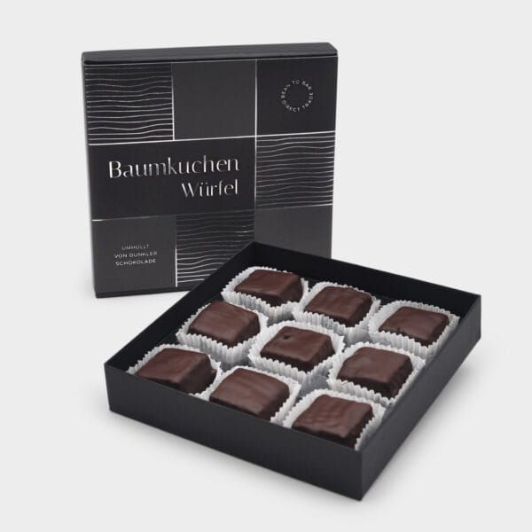 schwarze quadratische Geschenkbox mit 9 Baumkuchen Würfeln im Vordergrund geöffnet, dahinter die aufgestellte Box mit Silber geprägter Banderole.