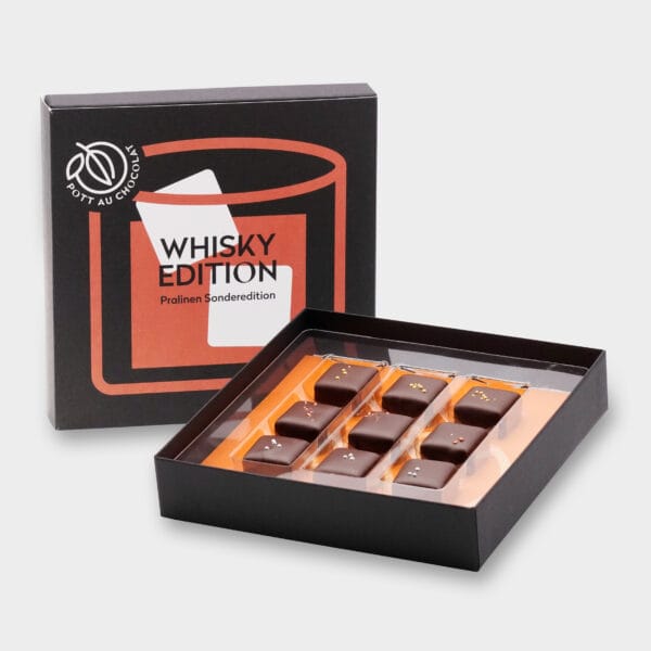 Aufgestellte Pralinenbox 'Whisky Edition'. Design in den Farben Schwarz, Orange und Weiß mit einer abstrakten Darstellung eines Glases mit Whisky und Eiswürfeln. Davor die geöffnete Box mit 9 Whisky-Pralinen in drei Reihen