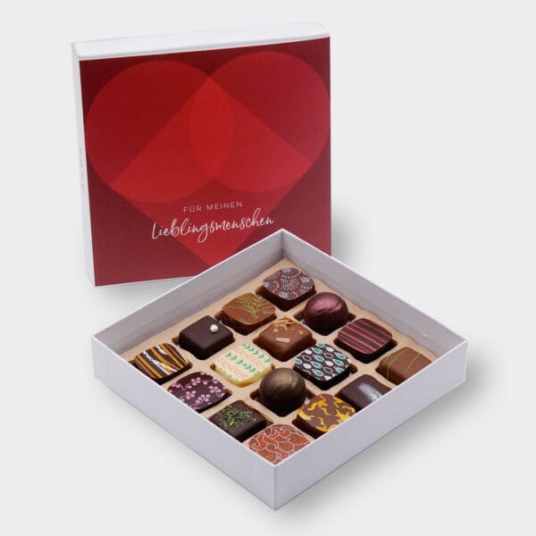 Pott au Chocolat weisse Pralinenbox mit 16 Pralinen und einer roten Banderole mit stilisiertem Herz und er Aufschrift "Für meinen Liebingsmenschen". Im Vodergrund die geöffnete Box. Im Hintergrund die Box mit Banderole aufrecht stehend
