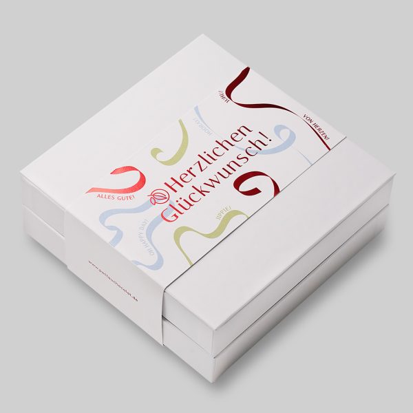 Doppelstöckige quadratische Pralinen Schachtel mit 'Herzlichen Glückwunsch' Motiv mit Luftschlangen in rot, blau und grün. Der Hintergrund des Motivs ist weiß, die rote Schrift ist geprägt und schimmert metallisch.