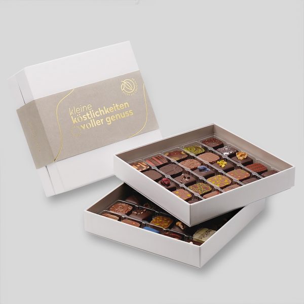 Weiße Pralinenbox mit grauer Banderole und goldener Schrift 'Kleine Köstlichkeiten voller Genuss'. Davor steht die Box geöffnet - 2 Ebenen mit jeweils 25 Pralinen.